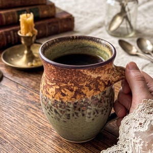 May include: A ceramic mug filled with dark liquid, likely coffee, with a textured brown, tan, and green exterior. The mug is held by a person. The background includes antique books, a candle, and silverware, suggesting a vintage aesthetic.