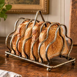 May include: A metal toast rack with a tarnished brass finish featuring a central handle and four curved slots holding four slices of golden-brown toasted bread. The rack stands on four small ball feet and has a wavy, scalloped wire frame design.