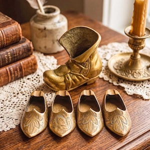 May include: A collection of antique brass decorative items, including a boot-shaped container and four small shoe-shaped ashtrays with leaf designs. A candle in a brass holder and a stack of old books are also visible.