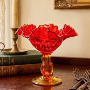 May include: A vibrant red glass bowl with a ruffled rim, set on a pedestal base. The bowl has a textured surface and a yellow-orange base. A candle and books are in the background. A decorative piece for display.