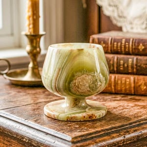 May include: A green and brown onyx goblet with a wide bowl and a pedestal base. The goblet is displayed on a wooden surface, with a brass candle holder and books in the background. The goblet's design is reminiscent of antique glassware.