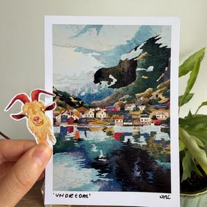 May include: A rectangular art print featuring a landscape of a coastal village with colourful houses, mountains, and water reflections, with the text 'UNDREOM' and 'NNC' at the bottom, accompanied by a separate die-cut sticker of a brown goat.