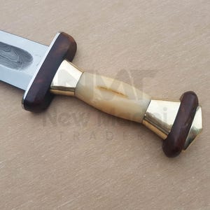 May include: A close-up of a dagger with a wooden handle and a brass pommel. The blade is made of Damascus steel and has a wavy pattern.