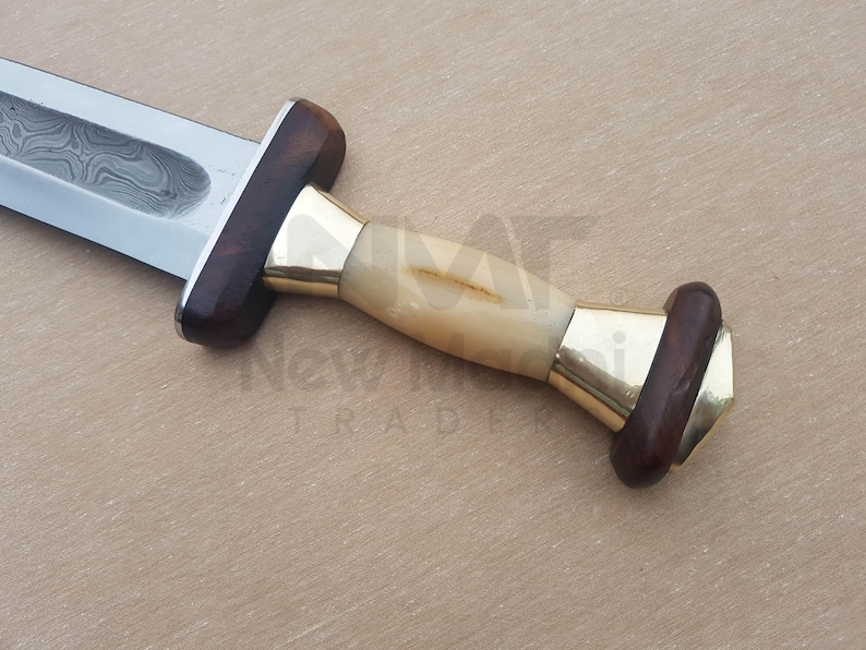 May include: A close-up of a dagger with a wooden handle and a brass pommel. The blade is made of Damascus steel and has a wavy pattern.