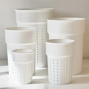 Op de afbeelding: Een set van vier witte, cilindrische kunststof houders van verschillende hoogtes, elk voorzien van een gladde bovenrand en een onderste gedeelte met verticale, rechthoekige sleuven in een rasterpatroon voor drainage.
