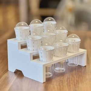 Op de afbeelding: Een witte, trapsgewijze displaystandaard met meerdere kleine, witte containers met doorzichtige, gewelfde deksels. De containers zijn in een trapsgewijs patroon gerangschikt, met doorzichtige bekers eronder. De display staat op een houten oppervlak.