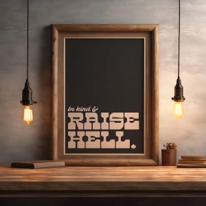 Puede incluir: Una impresión artística enmarcada con el texto "be kind & RAISE HELL" en una fuente retro. La obra de arte está en un marco de madera, sobre un fondo oscuro. La imagen está iluminada por dos bombillas colgantes, creando un ambiente cálido.
