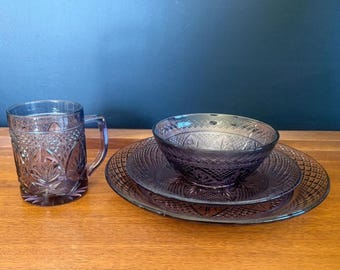 May include: A set of purple pressed glass dinnerware featuring a mug, a small bowl, and two stacked plates. Each piece displays a detailed diamond and starburst pattern across the surface, reflecting light through the translucent purple glass.