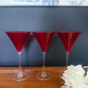 May include: Three martini glasses with deep red, conical bowls and clear, slender stems that transition into circular bases. The glasses feature a smooth, translucent finish on the red glass, providing a consistent color profile across all three items.