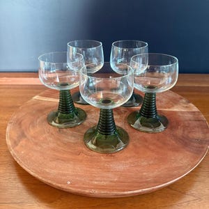 May include: Set of six clear glass wine glasses with green stems. The glasses are arranged on a round wooden tray. The stems have a ribbed design. The background is a dark blue wall.