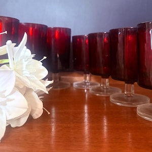 May include: A set of seven deep red glass goblets with clear stems and circular bases, arranged in a row. A cluster of white artificial flowers with layered petals is positioned in the foreground, partially obscuring the first red glass goblet.