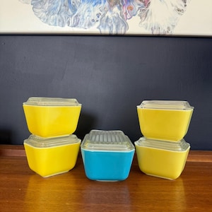 May include: A collection of vintage Pyrex refrigerator dishes. The set includes yellow and turquoise rectangular containers with clear, ridged lids. The containers are arranged on a wooden surface, showcasing their bright colors and classic design.