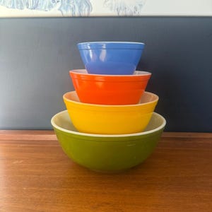 May include: A stack of four vintage mixing bowls in vibrant colors: blue, orange, yellow, and green. Each bowl has a white interior. The bowls are arranged from smallest to largest, creating a colorful display.