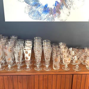 May include: A collection of vintage glassware, including champagne flutes, cocktail glasses, and tumblers, arranged on a wooden cabinet. Some glasses feature gold leaf designs, while others have etched patterns. The glassware is displayed in a well-lit setting.