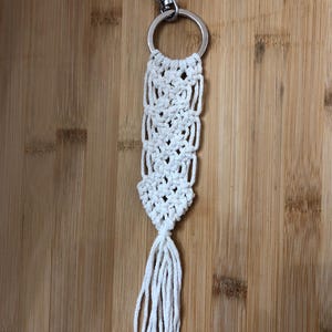 Op de afbeelding: Een handgemaakte macramé sleutelhanger in gebroken wit. De sleutelhanger heeft een geweven ontwerp met een zilverkleurige metalen ring en karabijnhaak. De onderkant van de sleutelhanger heeft een franje van losse koorden. De achtergrond is een houten oppervlak.