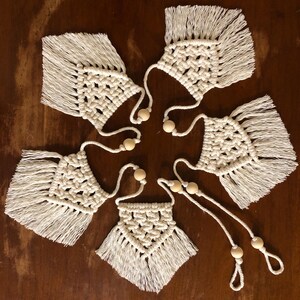 May include: Five white macrame wall hangings with wooden beads and fringe. The hangings are arranged in a circle on a wooden surface.