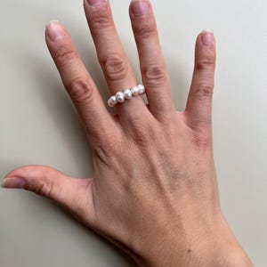 Könnte beinhalten: Ein Ring aus weißen Perlen wird an einer Hand getragen. Der Ring besteht aus mehreren runden, weißen Perlen, die aneinandergereiht sind. Die Hand ist vor einem neutralen Hintergrund positioniert, um das Design und die Farbe des Rings hervorzuheben.
