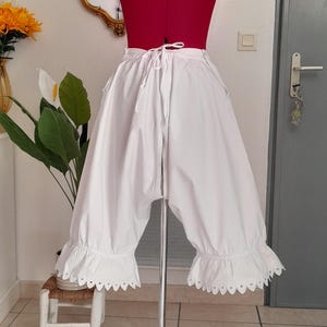 May include: White knee-length bloomers featuring a drawstring waist and ruffled leg openings with a decorative scalloped eyelet trim. The garment is displayed on a red mannequin torso, showcasing the loose, voluminous fit and gathered fabric details.