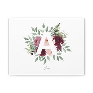Puede incluir: Impresión en lienzo con la letra "A" en blanco, rodeada de un diseño floral. Las flores son en tonos burdeos, malva y rosa, con follaje verde. El nombre "Ana" está escrito en cursiva debajo.