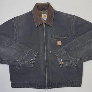 Puede incluir: Chaqueta de trabajo gris oscuro con cuello marr&oacute;n y cierre de cremallera. La chaqueta tiene un peque&ntilde;o bolsillo en el pecho y un parche con el logotipo. Las mangas tienen pu&ntilde;os abotonados. La chaqueta est&aacute; hecha de un material duradero.