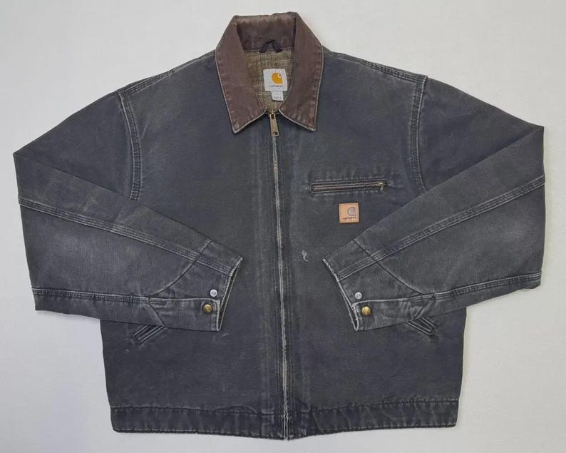 Puede incluir: Chaqueta de trabajo gris oscuro con cuello marr&oacute;n y cierre de cremallera. La chaqueta tiene un peque&ntilde;o bolsillo en el pecho y un parche con el logotipo. Las mangas tienen pu&ntilde;os abotonados. La chaqueta est&aacute; hecha de un material duradero.