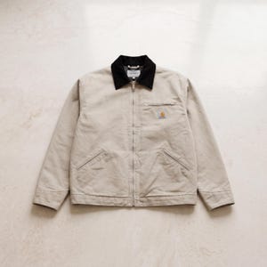 May include: A beige work jacket with a black collar and a zip closure. The jacket features two front pockets and a small logo patch on the chest. It is made of a durable material.