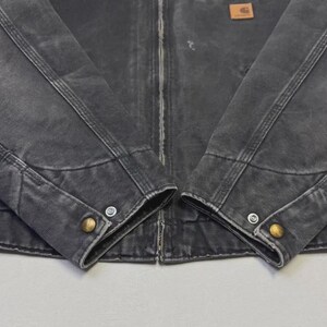 Puede incluir: Primer plano de una chaqueta desgastada de color carb&oacute;n con costuras visibles y botones color lat&oacute;n en los pu&ntilde;os. Un peque&ntilde;o parche rectangular marr&oacute;n est&aacute; cosido en la parte superior del pecho. La chaqueta tiene cierre de cremallera.