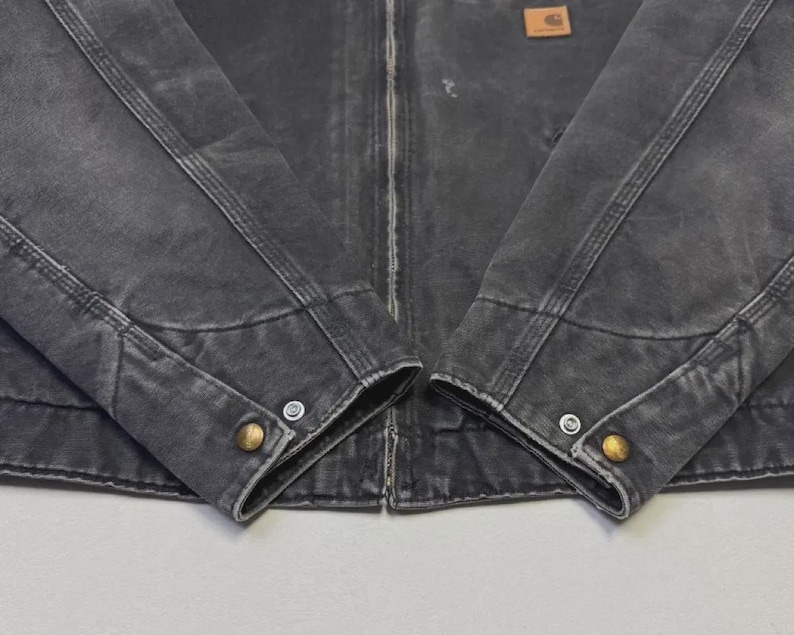 Puede incluir: Primer plano de una chaqueta desgastada de color carb&oacute;n con costuras visibles y botones color lat&oacute;n en los pu&ntilde;os. Un peque&ntilde;o parche rectangular marr&oacute;n est&aacute; cosido en la parte superior del pecho. La chaqueta tiene cierre de cremallera.