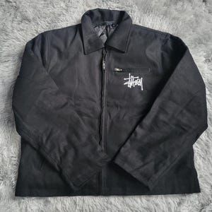 May include: Black jacket with a collar and a zip fastening. The jacket has a white logo on the chest and a small pocket. The jacket is made of a durable material, suitable for various weather conditions.