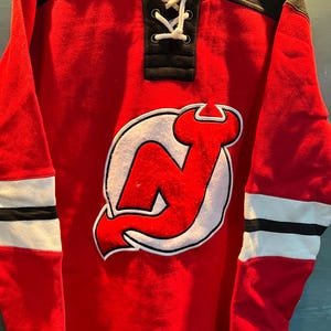 May include: Red hockey-style sweatshirt with the New Jersey Devils logo on the front. The sweatshirt has a black collar and a lace-up neckline. White and black stripes adorn the sleeves.