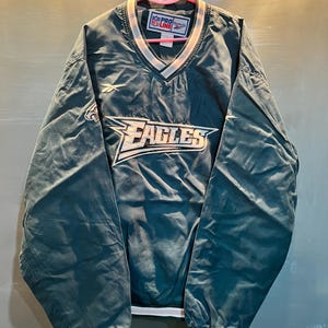 Puede incluir: Chaqueta tipo jersey verde azulado y blanca con la palabra "EAGLES" en el pecho. La chaqueta tiene cuello en V y ribetes blancos y rosas. El logotipo de los Eagles está en el pecho izquierdo. La chaqueta está colgada de una percha rosa.