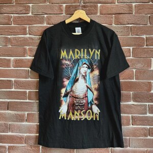 May include: Black t-shirt featuring a graphic of Marilyn Manson with the text "MARILYN" above and "MANSON" below the image. The image depicts a figure in a blue head covering and ornate clothing. The shirt is hanging on a wooden hanger.