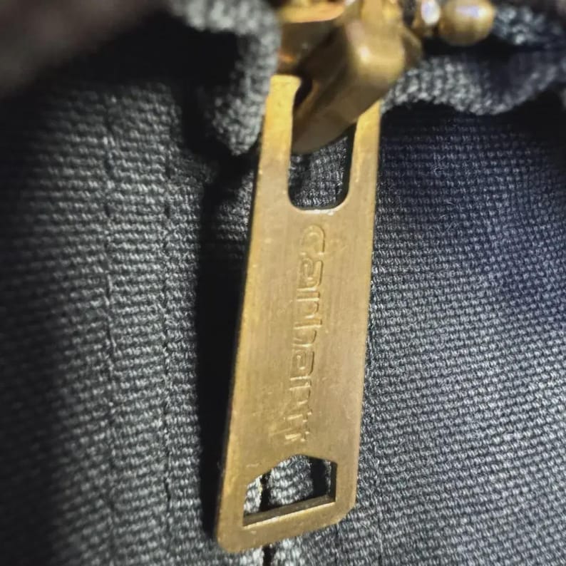 Puede incluir: Primer plano de un tirador de cremallera de lat&oacute;n con el nombre de la marca "Carhartt" en relieve. La cremallera est&aacute; sujeta a una tela gris oscuro, posiblemente mezclilla. La cremallera est&aacute; parcialmente cerrada.