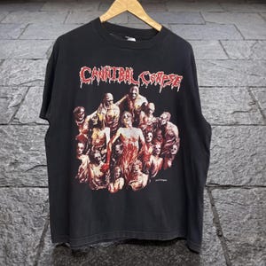 May include: Black t-shirt featuring the band name "Cannibal Corpse" in red, stylized lettering above a graphic of several figures with exposed flesh. The figures are in shades of red, white, and brown.
