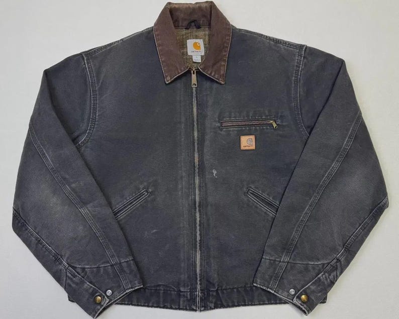 Puede incluir: Chaqueta Carhartt gris oscuro con cuello marr&oacute;n y cremallera completa. La chaqueta tiene dos bolsillos delanteros, un bolsillo en el pecho con cremallera y un peque&ntilde;o parche con el logotipo. La chaqueta est&aacute; hecha de un material duradero y desgastado.