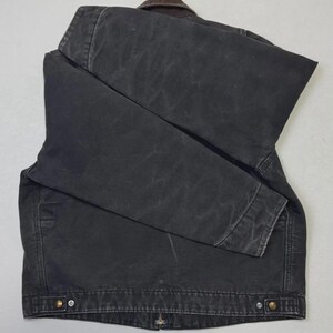 Puede incluir: Chaqueta gris oscuro con cuello marr&oacute;n, doblada sobre una superficie gris claro. La chaqueta tiene cierre de botones y dos bolsillos delanteros. La tela parece ser un material duradero y desgastado.