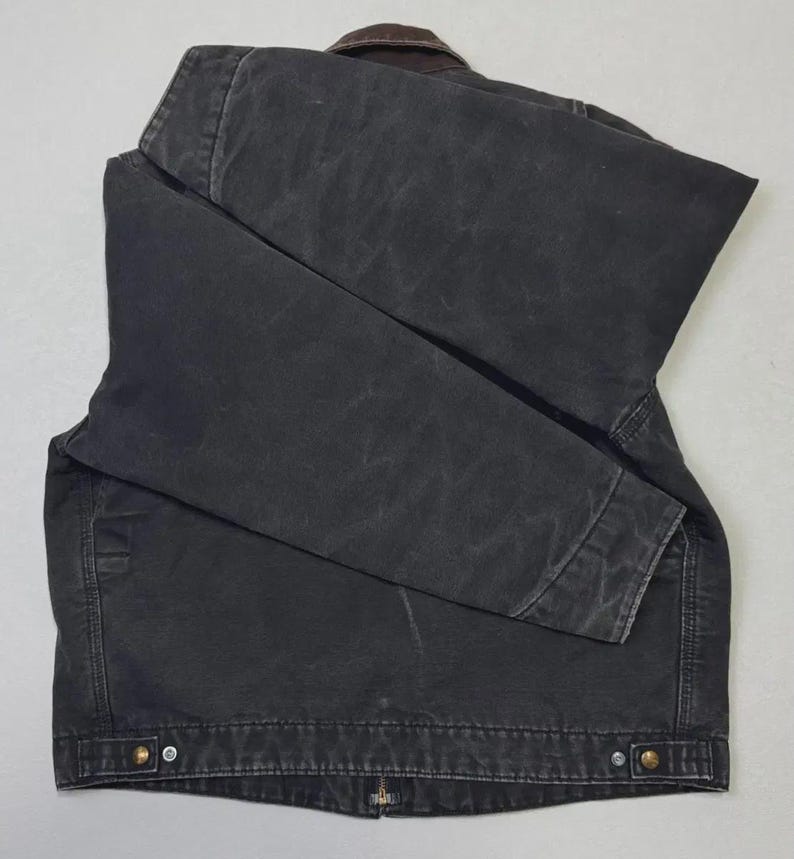 Puede incluir: Chaqueta gris oscuro con cuello marr&oacute;n, doblada sobre una superficie gris claro. La chaqueta tiene cierre de botones y dos bolsillos delanteros. La tela parece ser un material duradero y desgastado.