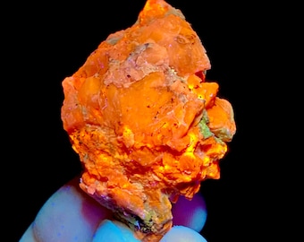 May include: A raw, crystalline mineral specimen exhibiting intense fluorescent orange coloration. The rock features a jagged, uneven surface with multiple facets and crystalline structures that glow brightly against a dark background.