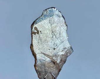 May include: A translucent, irregularly shaped, gray-toned mineral specimen. The stone has a layered appearance with visible internal structures and is held by a silver-colored metal clamp.