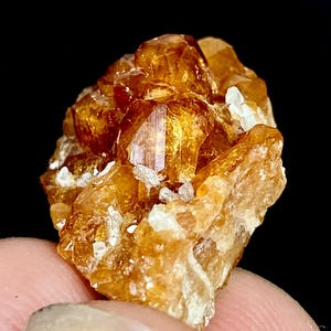 Pode incluir: Close-up de um aglomerado de cristais laranja-dourado com estrutura cristalina. Os cristais têm uma aparência brilhante e são acentuados por pequenas formações brancas irregulares. O fundo é preto, destacando a cor e a textura do mineral.