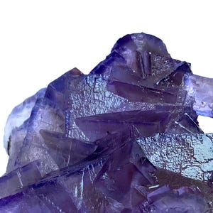 Könnte beinhalten: Nahaufnahme eines violetten Fluorit-Kristallclusters. Der Kristall hat eine geschichtete, geometrische Struktur mit tiefvioletter Farbe und strukturierter Oberfläche. Der Hintergrund ist weiß.