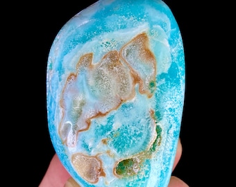 May include: A polished, rounded gemstone featuring a vibrant turquoise blue base color with intricate, swirling patterns of white and earthy brown mineral inclusions. The stone has a smooth, glossy surface texture and an irregular, organic shape.