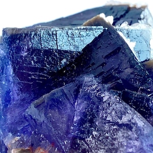 Puede incluir: Primer plano de un grupo de cristales de fluorita. El cristal presenta una paleta de colores azul oscuro y púrpura con una estructura cristalina. Las facetas del cristal reflejan la luz, creando un efecto brillante. La apariencia general es la de un espécimen mineral natural.