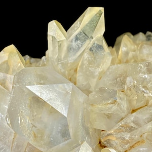 May include: A cluster of translucent, pale yellow quartz crystals with sharp, pointed terminations and multifaceted surfaces. The mineral formation features varying sizes of crystalline structures that catch the light, creating a bright, reflective appearance against a dark background.