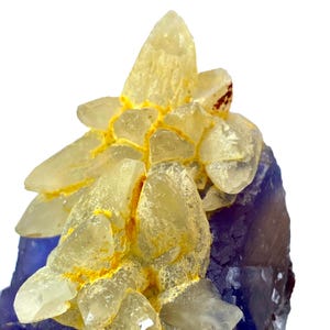 Könnte beinhalten: Ein Mineralexemplar mit einer Ansammlung durchscheinender, blassgelber Kristalle auf einer tiefvioletten Basis. Die Kristallformation zeigt scharfe, kantige Spitzen, wobei leuchtend gelbe Mineralablagerungen die Zwischenräume ausfüllen.