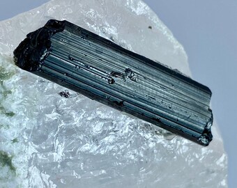 May include: A long, slender, dark green to black tourmaline crystal with prominent vertical striations resting on a translucent, jagged white quartz matrix. The mineral specimen displays a high-gloss finish and a distinct prismatic shape.
