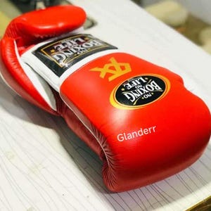 K&ouml;nnte beinhalten: Rote Boxhandschuhe mit wei&szlig;em Besatz und dem Text "No Life No Boxing" in einem goldenen Kreis. Die Handschuhe haben ein wei&szlig;es Logo mit einem schwarzen "X" und dem Text "Glanderr" in wei&szlig;.