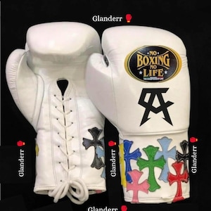 May include: Pair of white boxing gloves with black stitching and colorful cross designs on the wrist area. The gloves have a black and gold patch with the text "NO BOXING NO LIFE".