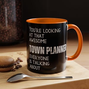 May include: Black coffee mug with an orange rim. The mug has white text that reads "You're looking at that awesome Town Planner everyone is talking about."
