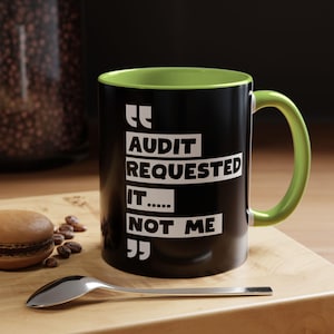 May include: Black coffee mug with a green interior and handle. The mug has the text "AUDIT REQUESTED IT..... NOT ME" in white. A silver spoon and a macaron are on a wooden surface.